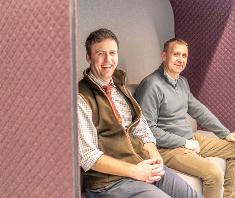 Huw and Iolo sit side by side on a cushioned bench in a modern lounge area with purple quilted walls, both looking toward the camera and smiling - discussing the latest rural property to let in Wales.