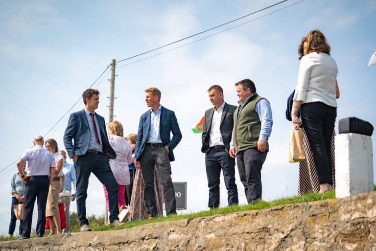 A group of people, mostly men in suits, stand and talk outdoors on a sunny day near a low stone wall with a utility pole and wires in the background, possibly discussing plans as land and property consultants.