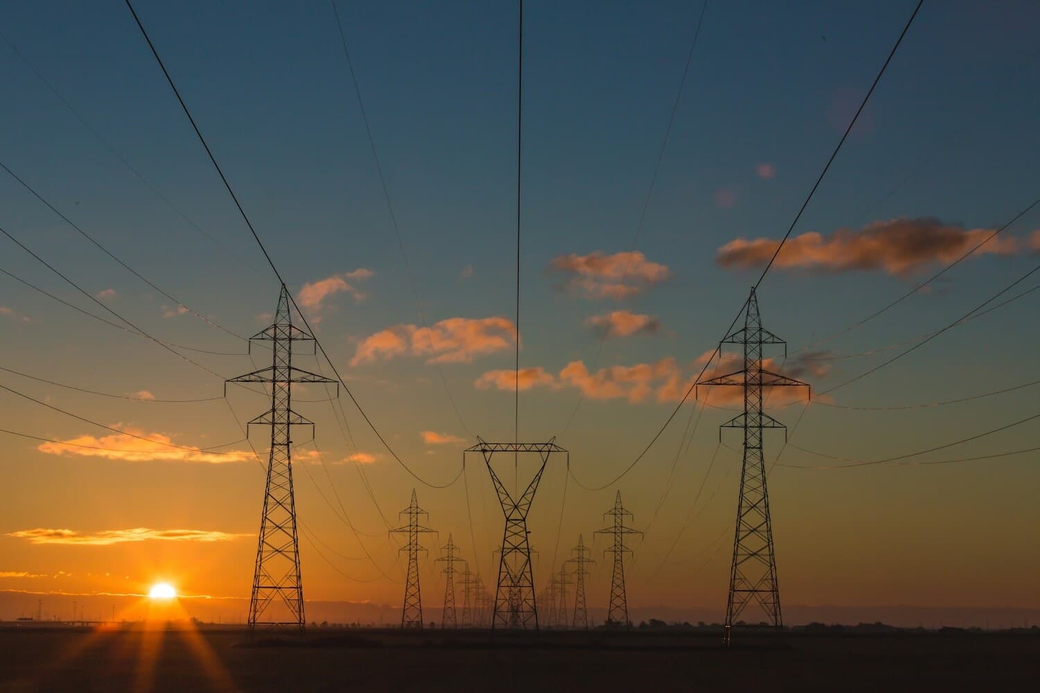 High-voltage power lines and transmission towers stretch across a flat landscape at sunset, symbolizing the progress of renewable energy development beneath a clear sky with scattered clouds.
