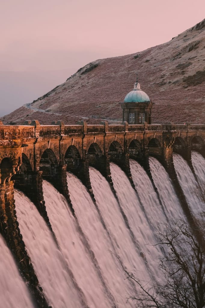 A stone dam with arched spillways releases water down its face. A small domed building sits above the dam, and grassy hills rise in the background under a pink sky.