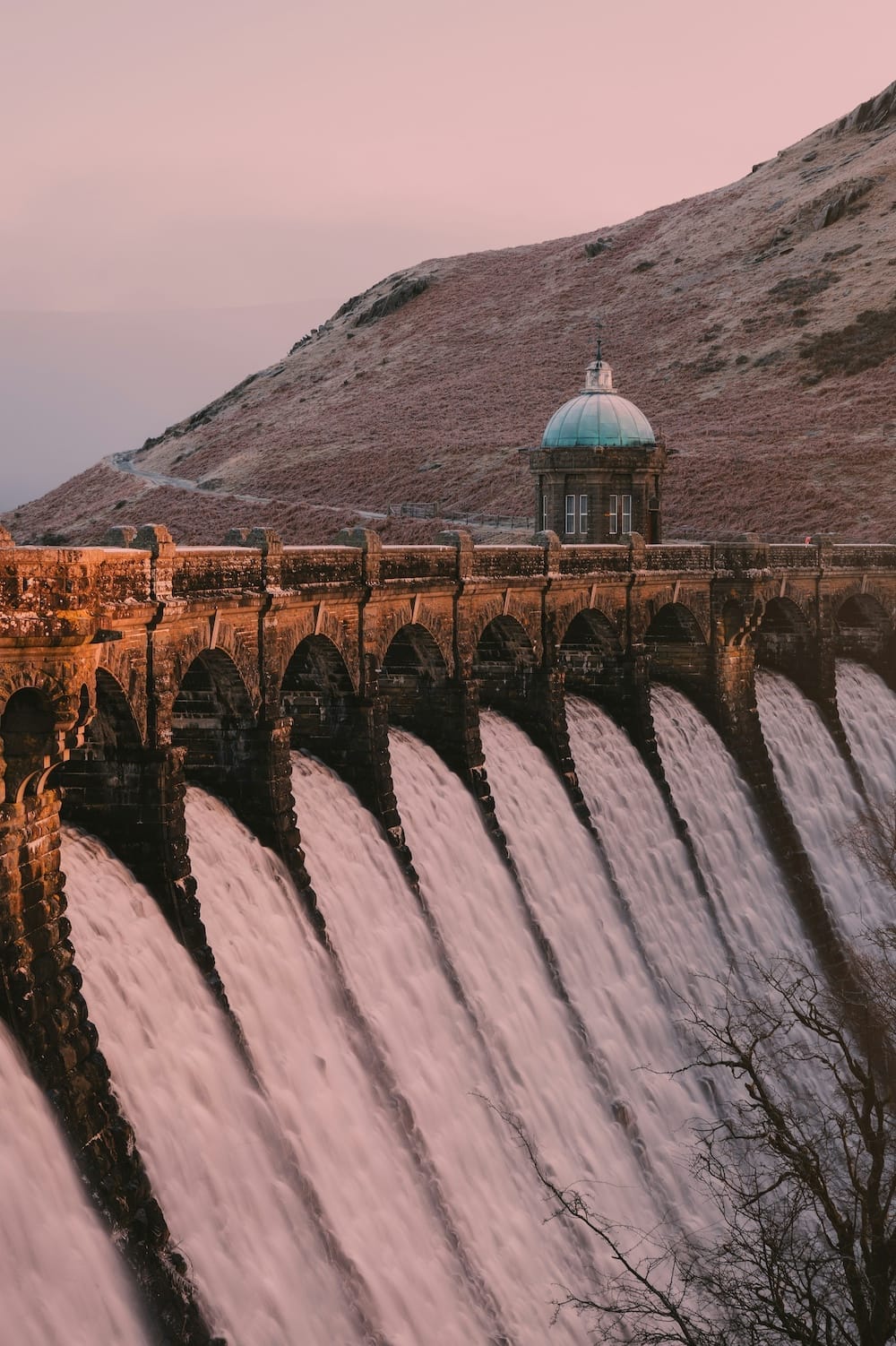 A stone dam with arched spillways releases water down its face. A small domed building sits above the dam, and grassy hills rise in the background under a pink sky.