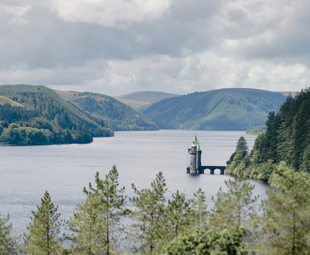 A stone tower with a green roof stands on a bridge in the middle of a lake, surrounded by forested hills and cloudy skies.