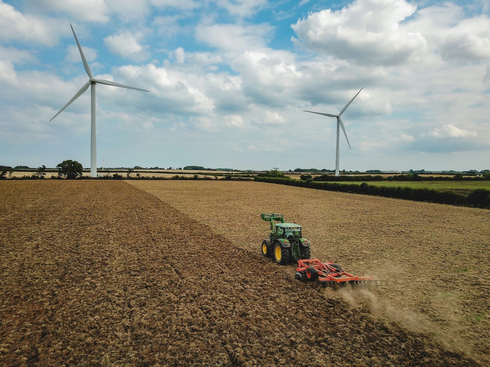 A tractor plows a large field under a partly cloudy sky, with two wind turbines standing in the background.