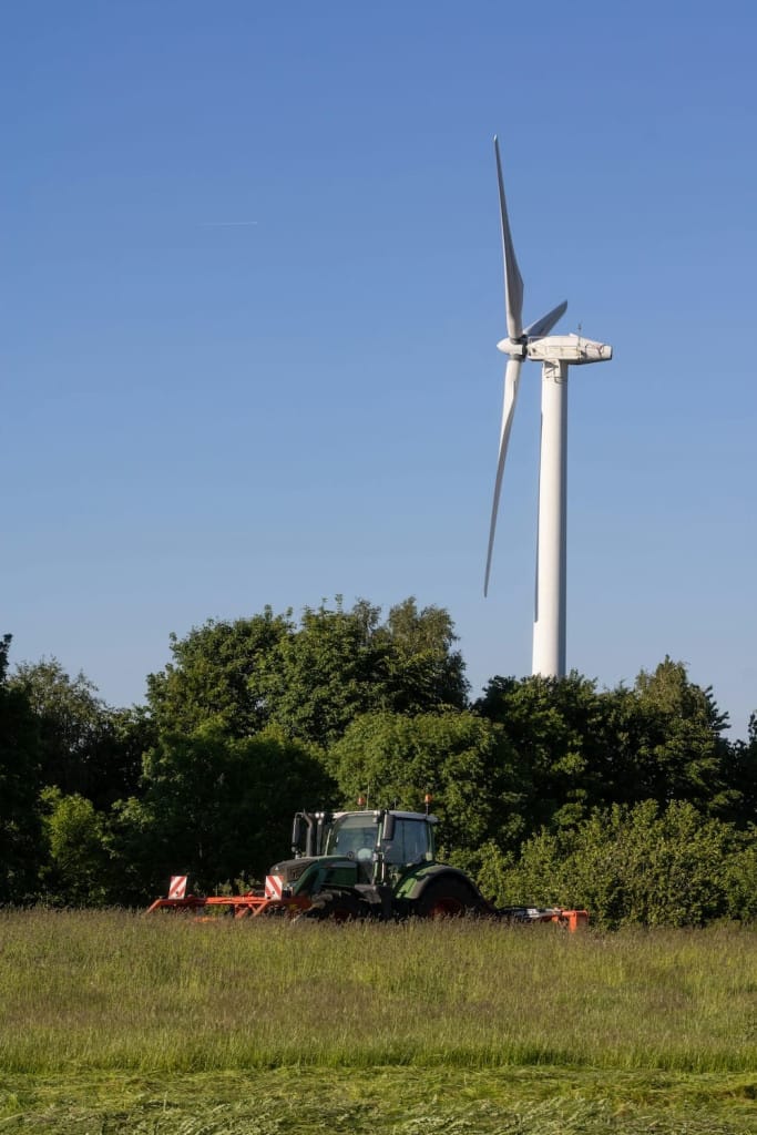 A tractor works in a grassy field with trees in the background and a wind turbine standing tall against a clear blue sky.