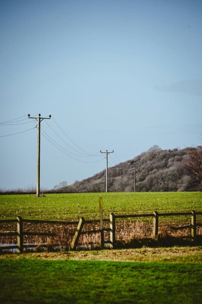 Wooden utility poles with power lines cross a grassy field, with a wooden fence in the foreground and a tree-covered hill in the background under a clear blue sky.