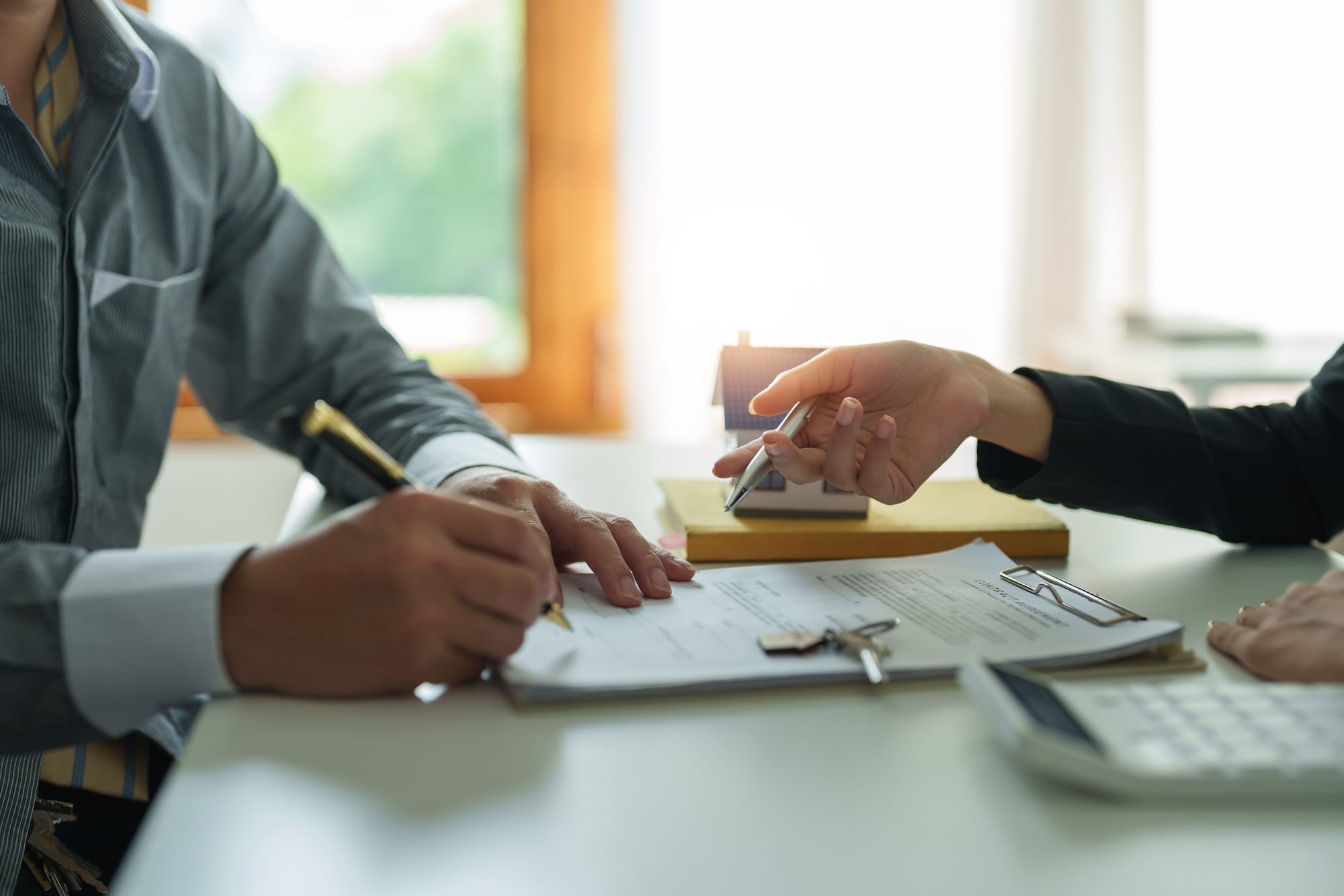 Two people sit at a table; one signs a document while the other points to it. A house model and keys rest nearby, suggesting a real estate or contract signing.