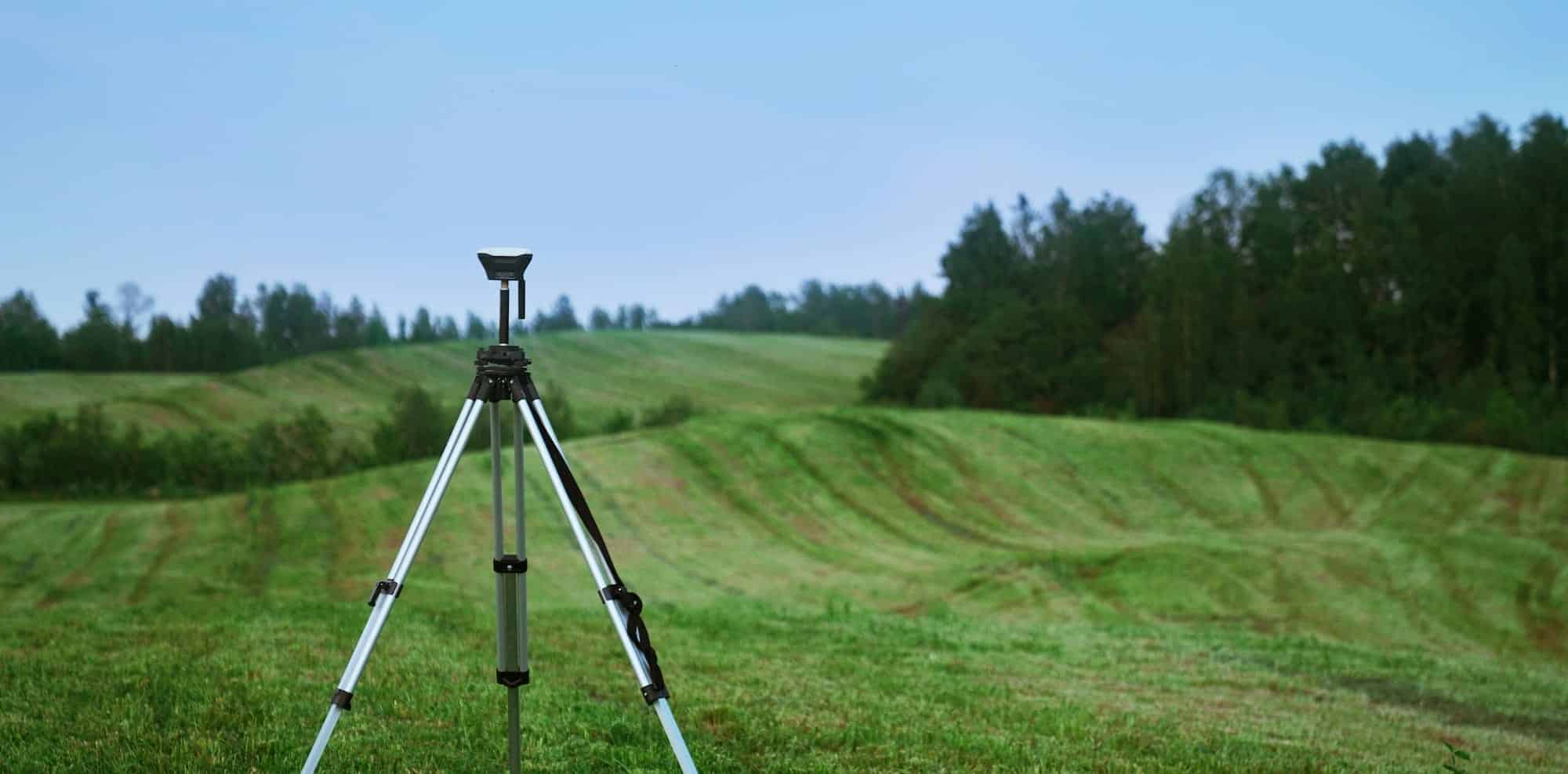 A tripod stands on a grassy field with rolling hills and a line of trees in the background under a clear blue sky.
