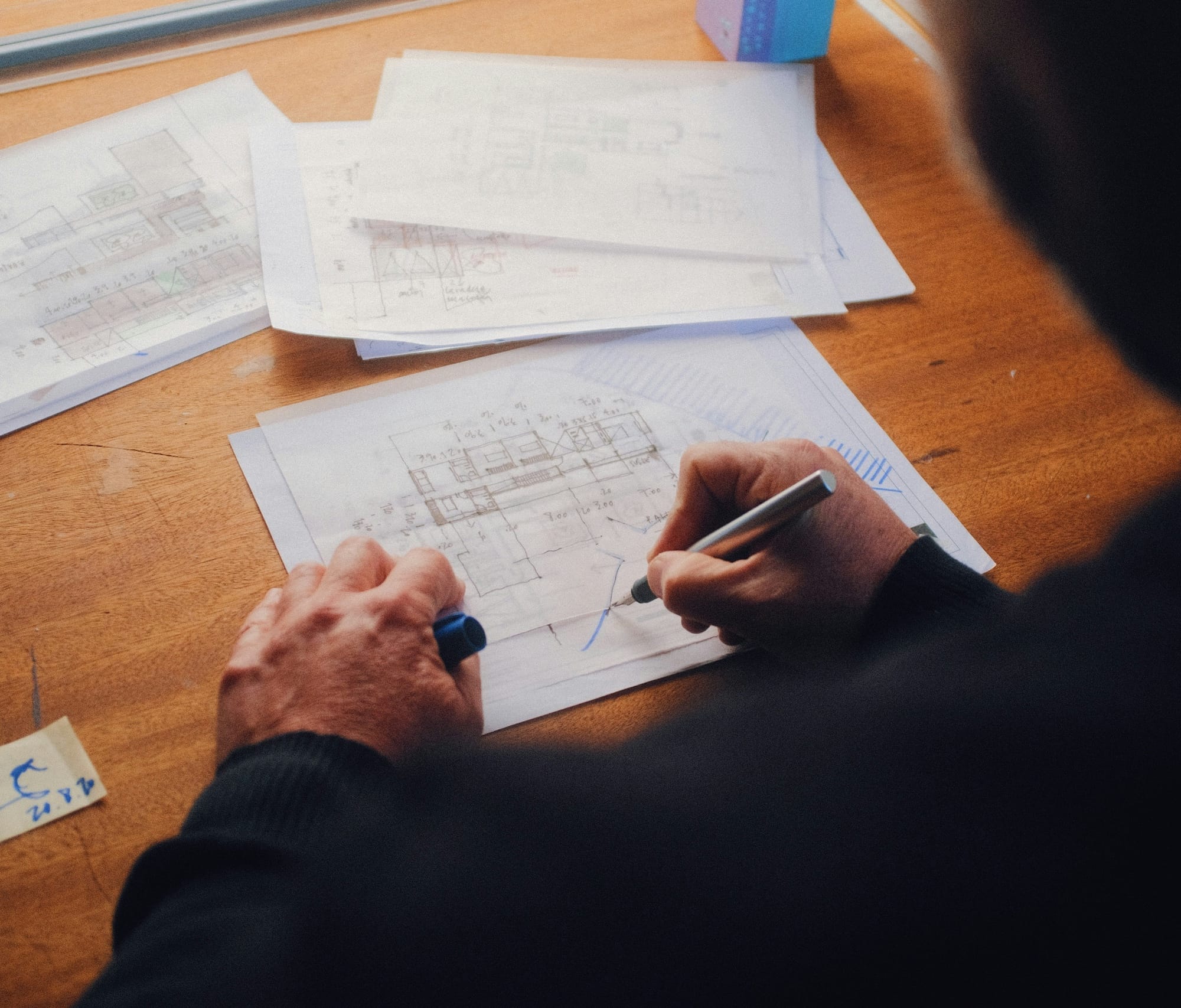 Person drawing architectural plans on paper at a wooden desk, surrounded by additional technical drawings and documents.