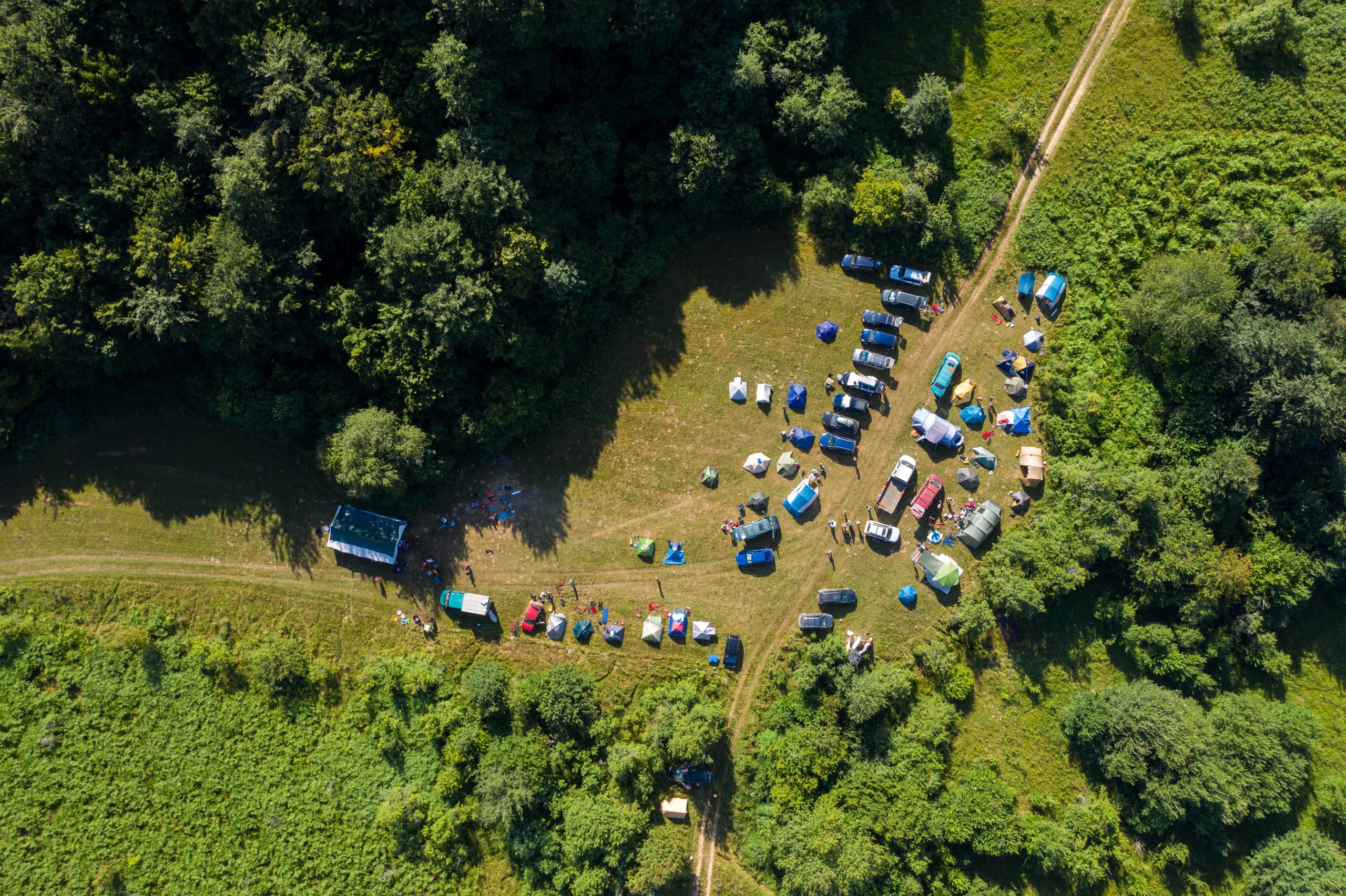 Aerial view of a campsite with tents, vehicles, and people scattered along a grassy clearing next to a forest. Dirt paths border the area and dense greenery surrounds the site.