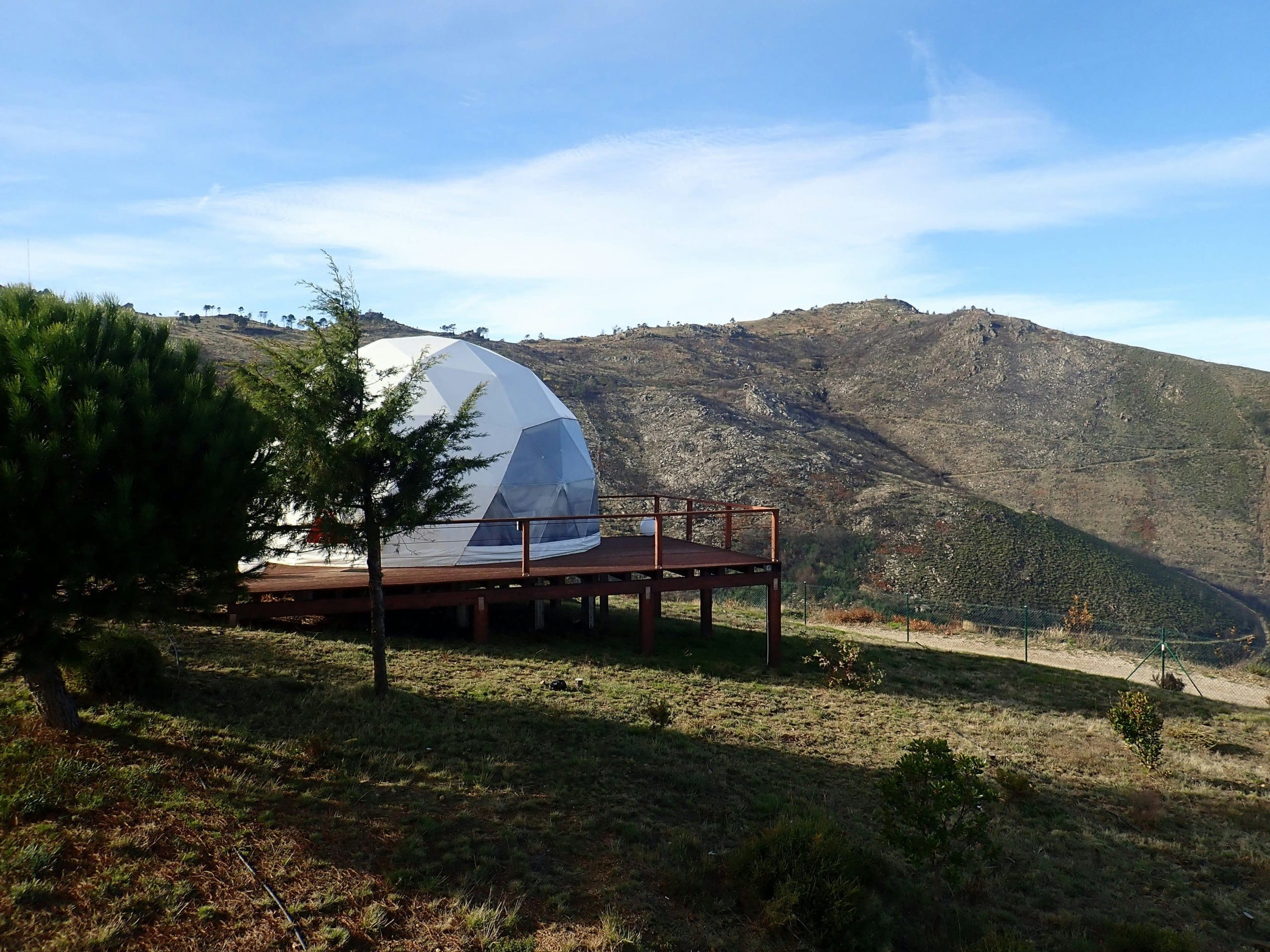 A white geodesic dome is situated on a wooden platform in a hilly, rural landscape with sparse vegetation and distant mountains under a clear blue sky.
