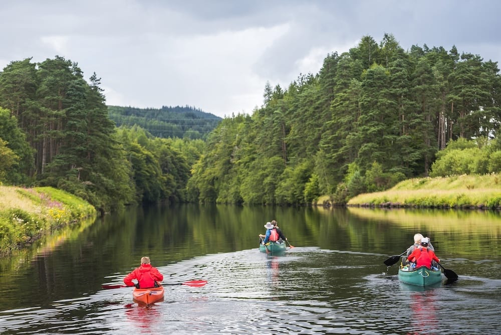 Three people paddle in two canoes and one kayak on a calm river surrounded by dense green trees under a cloudy sky.