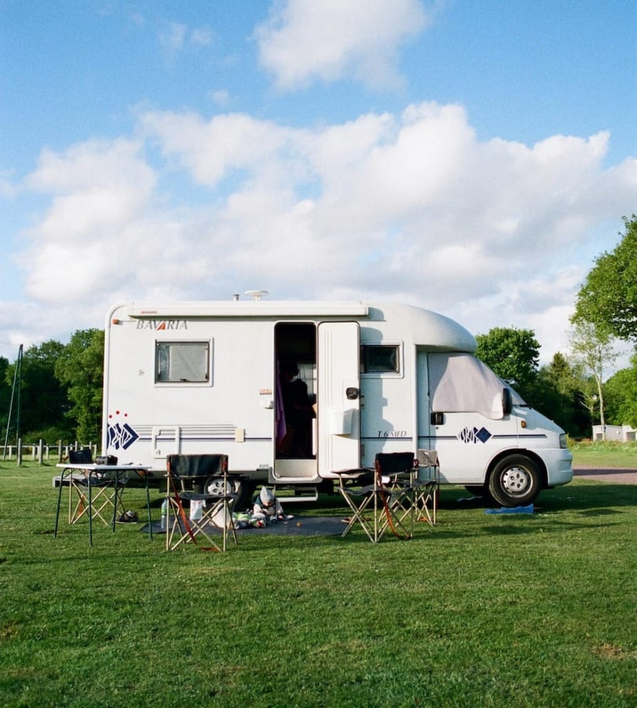 A white camper van is parked on grass with several folding chairs and a small table set up outside under a partly cloudy sky.