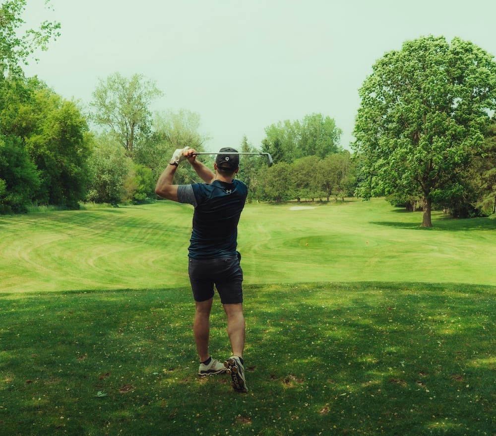 A person wearing a cap and shorts swings a golf club on a green golf course surrounded by trees on a clear day.