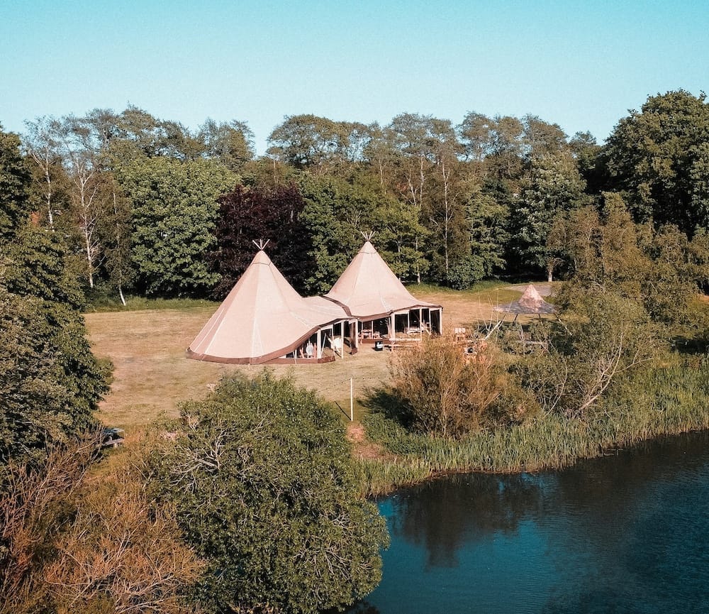 Two large tipi tents are set up on a grassy clearing near a pond, surrounded by trees under a clear blue sky.