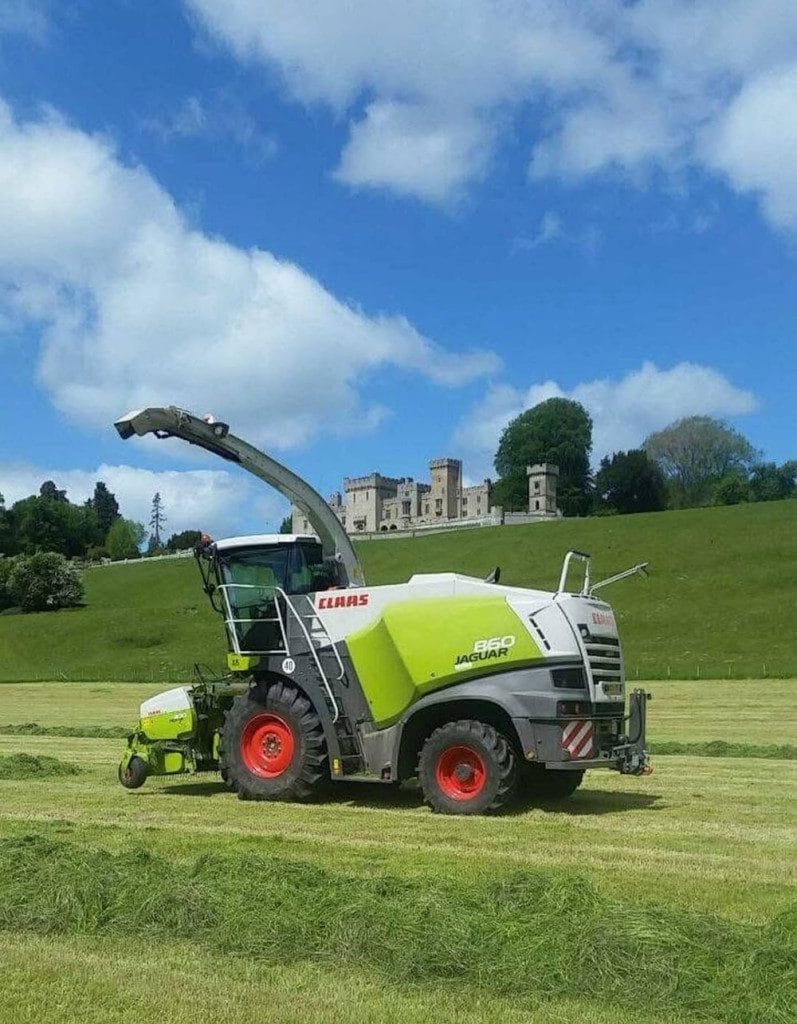 A CLAAS Jaguar forage harvester collects grass in a field with a castle on a hill in the background under a partly cloudy sky.