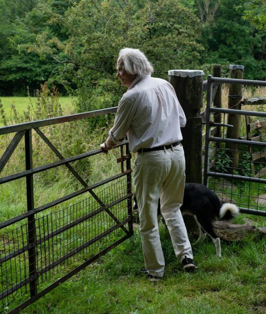 Older man in light clothing opens a metal gate in a grassy, rural area while a black and white dog stands beside him. Trees and fields are visible in the background.