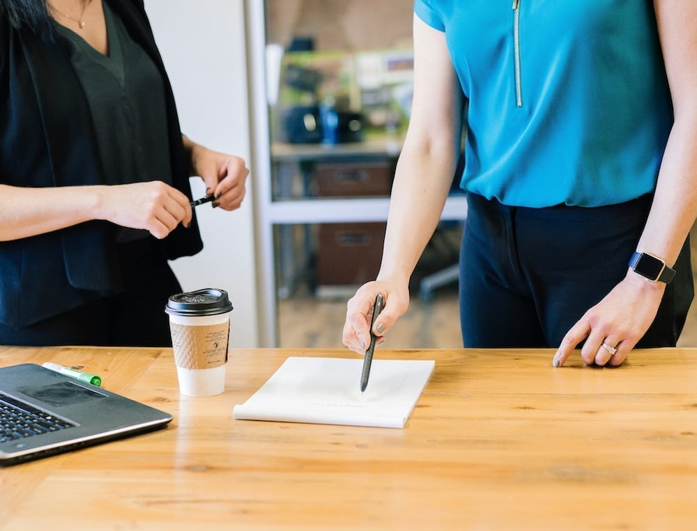 Two people stand at a wooden table with a notepad, pen, coffee cup, and laptop, engaged in a discussion that suggests a rural chartered surveyor meeting to review plans or property details.
