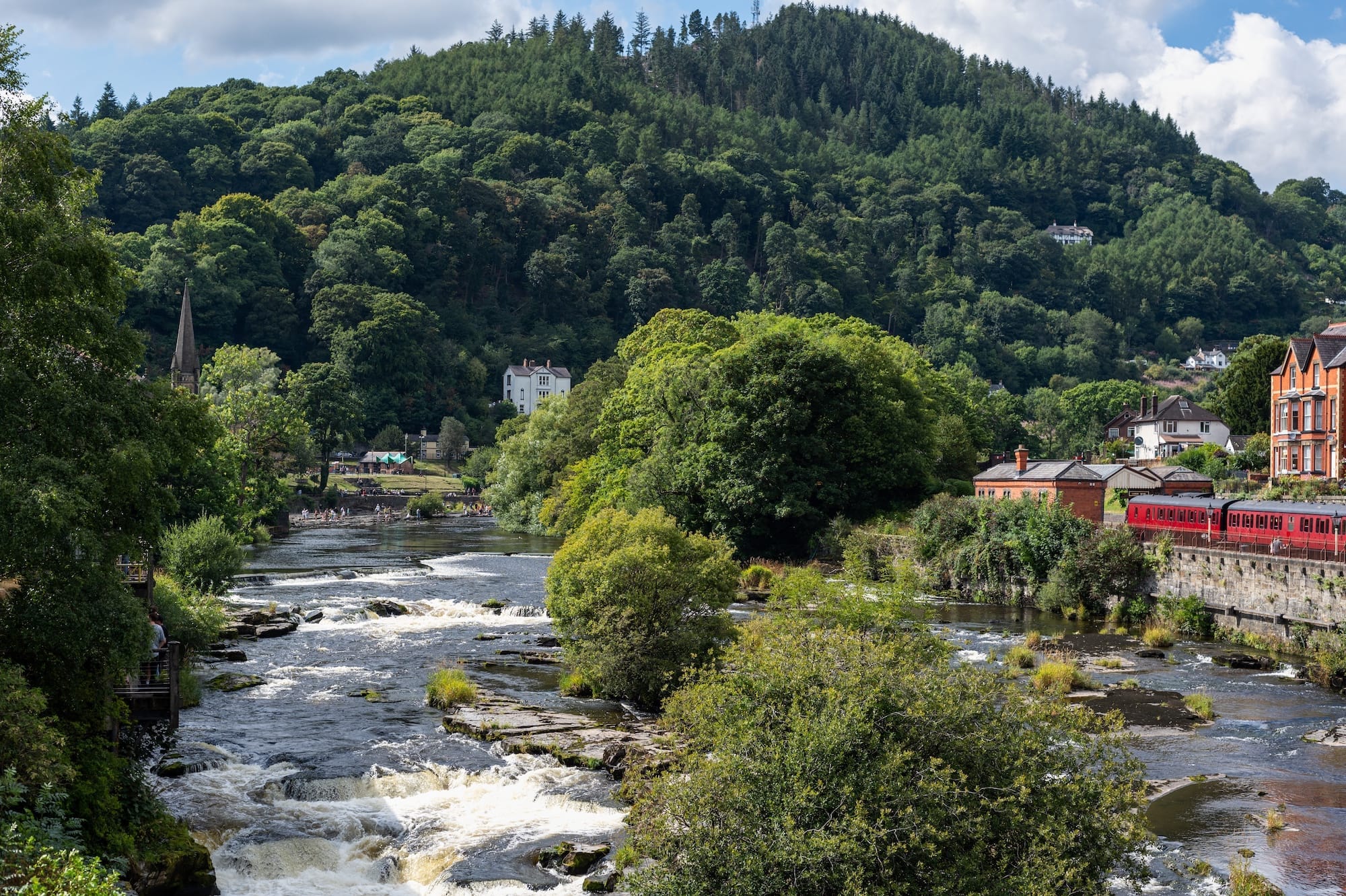 The Llangollen river flows past green trees and buildings, including a red train on the right, with forested hills in the background.