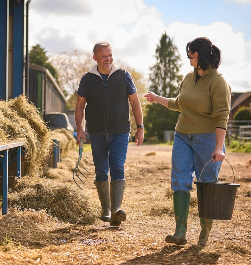 Two people in casual farm clothes and boots walk outside near hay; one carries a pitchfork and the other holds a bucket while they talk.