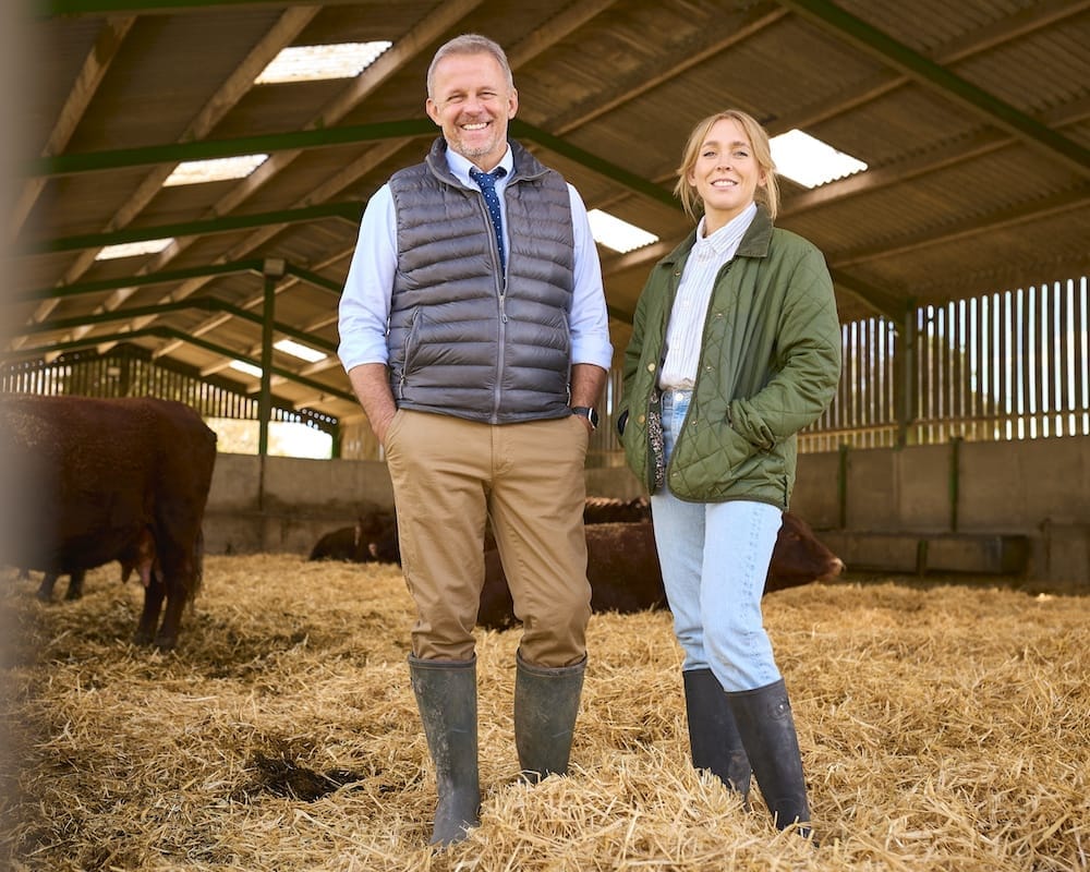 Two people wearing boots and farm clothing stand in a barn with hay on the ground and cows in the background.