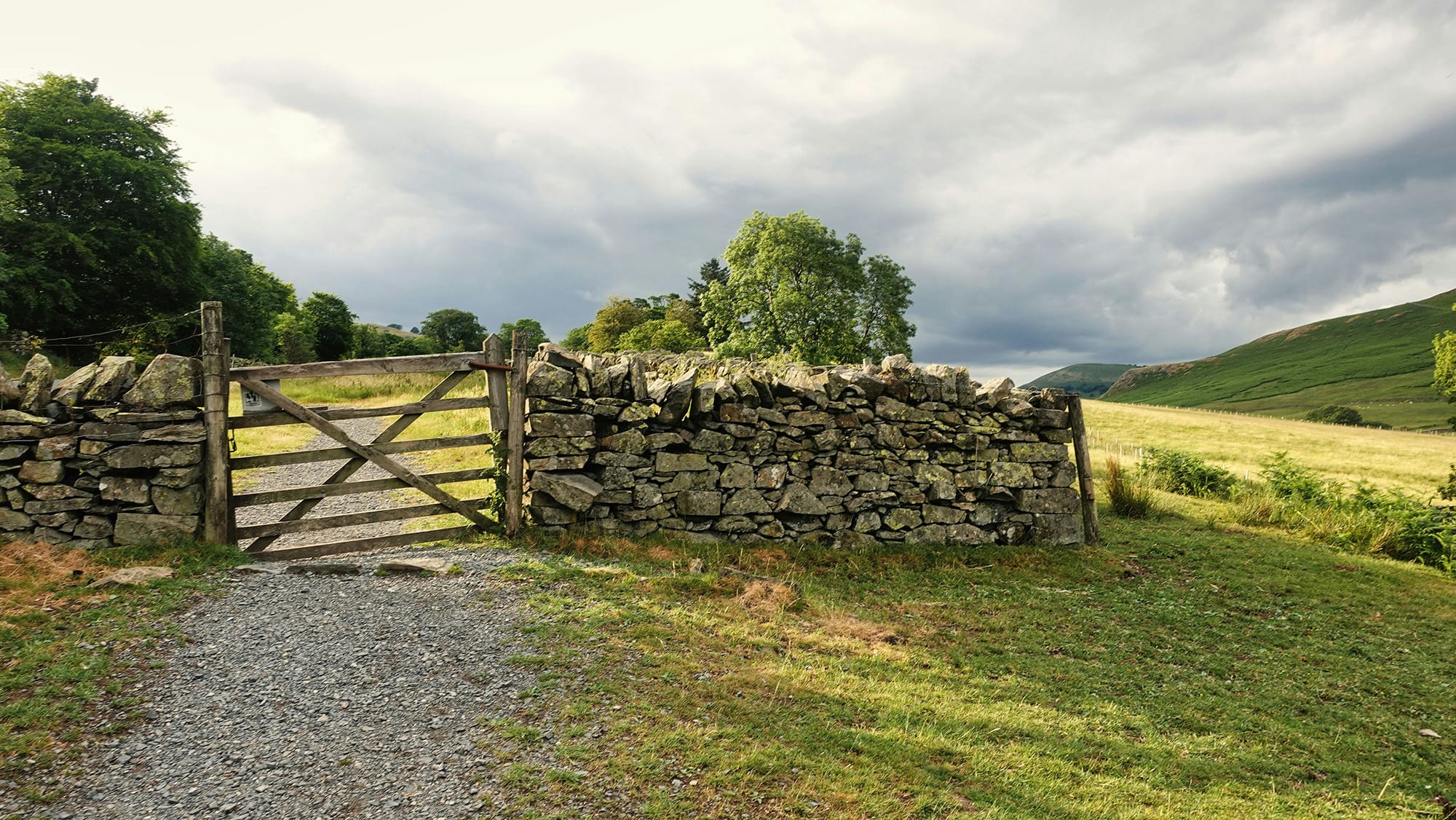 Mae giât bren wedi'i gosod mewn wal gerrig yn agor i gae glaswelltog, gyda llwybr graean yn arwain i fyny a bryniau tonnog o dan awyr gymylog yn y cefndir.