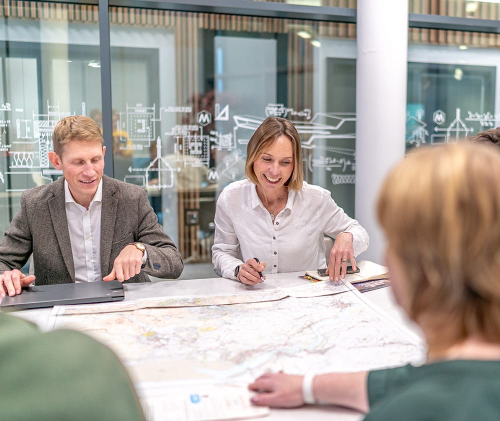 The Baileys team sit around a table examining a large map, with two of them smiling and discussing the contents; office environment in the background.