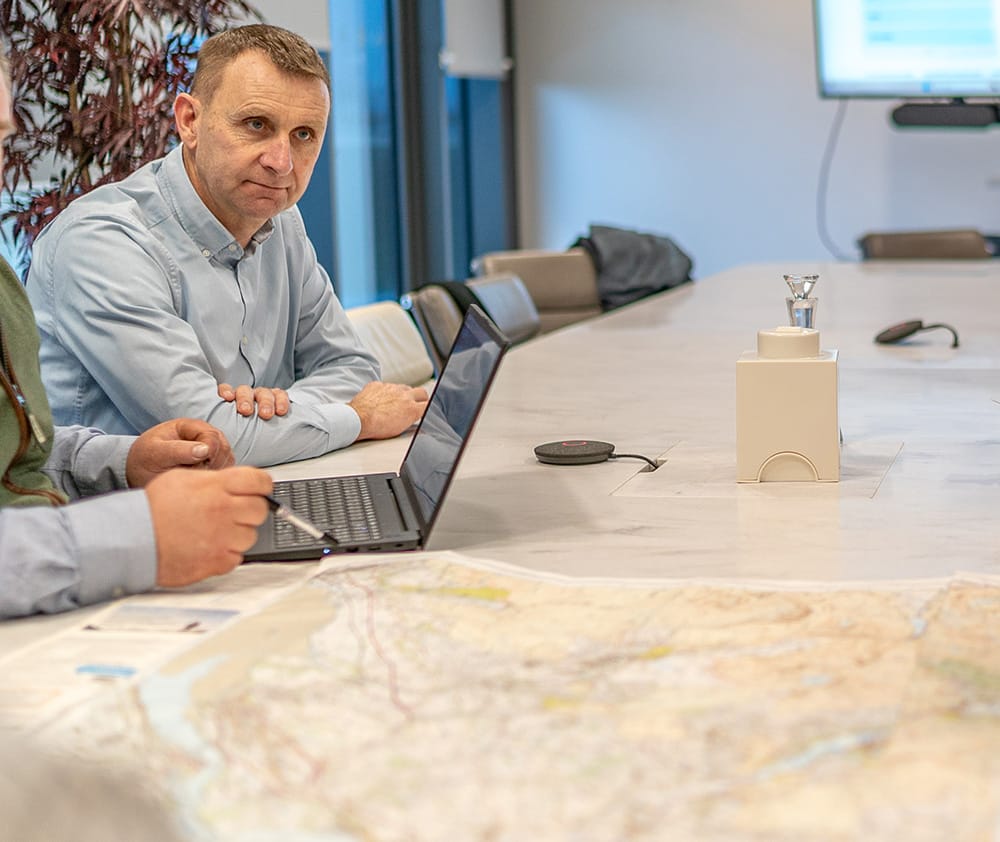 Iolo sits at a conference table with a large map and a laptop, engaged in discussion. A monitor is visible in the background.