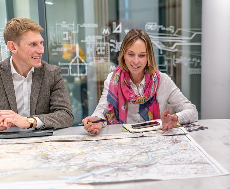 Ed and Helen sit at a table, discussing a large map spread out in front of them. Both have notebooks and pens, with diagrams visible on a glass wall in the background.