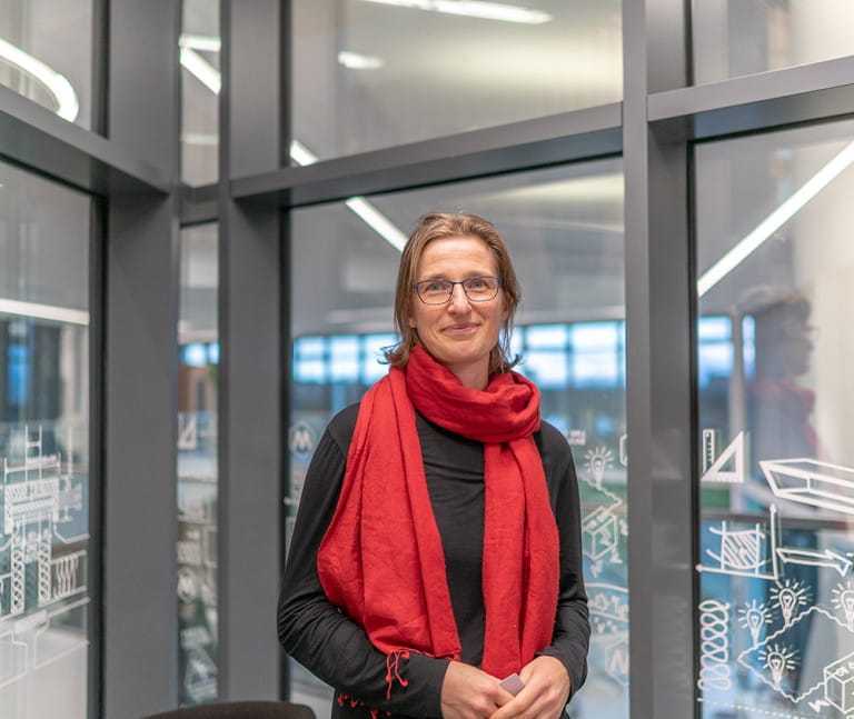 Jodie stands indoors near glass walls decorated with white geometric drawings.